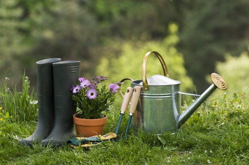 In a lush, well-maintained outdoor garden area, a pair of black rubber gardening boots stands upright on a vibrant green lawn near a small terracotta pot filled with blooming purple and white flowers. Nearby, there is an assortment of gardening tools including hand trowels and pruning shears, arranged on the grass. A silver metal watering can with a wooden handle and a long spout is positioned next to the tools, indicating ongoing garden care activities. The background features blurred trees and shrubbery, suggesting a spacious, landscaped garden environment typical of properties in Mayfair or similar upscale neighbourhoods. The scene is bathed in natural daylight, highlighting the rich textures of the grass, the glossy surface of the watering can, and the colorful flowers, creating a peaceful and inviting outdoor space suitable for regular garden and lawn maintenance, provided by companies like Lawn Mowing Mayfair.