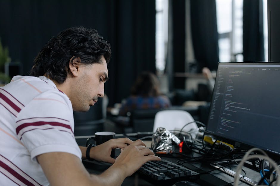 A young man with dark, wavy hair working intently at a desk in an office environment with natural light streaming through large windows. He is seated in front of a computer monitor displaying lines of